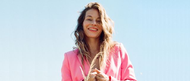 Portrait von Anna Rossinelli vor hellblauem Himmel. Sie lächelt selbstbewusst in die Kamera und trägt einen auffälligen pinken Blazer.