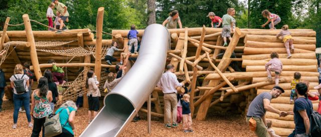 Ansicht Spielplatz im Hardwald, Viele Kinder spielen auf dem Holzspielplatz oder nutzen die Rutschbahn. Im Hintergrund sind grüne Bäume zu sehen.