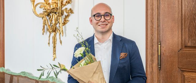 Porträt von Bürgerrat Marco Natoli nach der Wahl. Er hält einen Blumenstrauss in der Hand und steht in der Bel Etage des Stadthauses vor einer goldenen Wandleuchte und einer Holztüre.