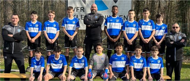 Mannschaftsfoto des Nachwuchses vom Fussballclub VfR Kleinhüningen in blau-weissen Trikots und schwarzen kurzen Hosen. Links, in der Mitte und rechts stehen die Trainer in schwarzen langen Trainingsanzügen. Juan Luis Gomez steht in der Mitte. Im Hintergrund ist die Club-Fahne zu sehen.