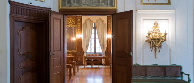 Ein prunkvoller Raum mit geöffneten Holztüren gibt den Blick frei auf ein reich verziertes Zimmer mit goldenen Wandverzierungen, Vorhängen und einem Kronleuchter. Vor den Türen steht eine elegante, grün gestrichene Holzbank mit Geflecht.