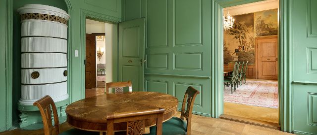 Ein eleganter Raum mit grünen Täferwänden, einem runden Holztisch und vier Stühlen im Biedermeier-Stil, daneben ein weisser Kachelofen in einer Wandnische. Durch zwei offene Türen sind weitere historisch eingerichtete Räume mit Wandgemälden, Kronleuchtern und antiken Möbeln sichtbar.