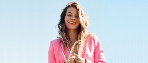 Portrait von Anna Rossinelli vor hellblauem Himmel. Sie lächelt selbstbewusst in die Kamera und trägt einen auffälligen pinken Blazer.