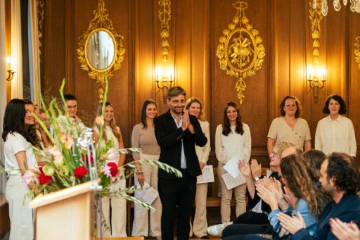 Der Dirigent des Basler Chors Les Voix steht im Anzug vor einem lächelnden Publikum und bedankt sich mit gefalteten Händen, während das Publikum applaudiert. Die Szene spielt in einem festlich dekorierten Raum im Stadthaus, dem Bürgergemeinderatssaal, er hat goldenen Verzierungen und ein Blumenarrangement steht in der linken Bildhälfte. 