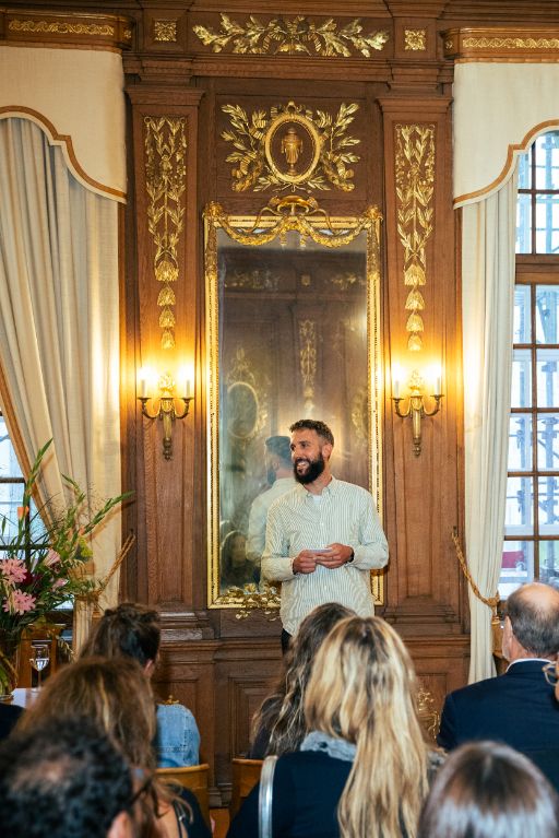 Der Dirigent des Basler Chors Les Voix steht im Anzug vor einem läAlain Schnetz vom Musikbüro Basel hält an der BG-Preisverleihung 2025 an Anna Rossinelli, im Bürgergemeinderatssaal die Laudatio vor dem Publikum.helnden Publikum und bedankt sich mit gefalteten Händen, während das Publikum applaudiert. Die Szene spielt in einem festlich dekorierten Raum im Stadthaus, dem Bürgergemeinderatssaal, er hat goldenen Verzierungen und ein Blumenarrangement steht in der linken Bildhälfte. 