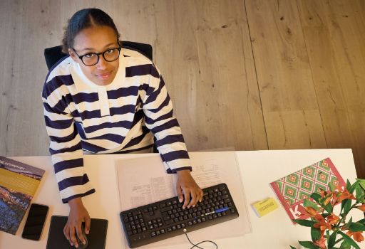 Eine Person mit Brille und gestreiftem Pullover sitzt an einem Schreibtisch, bedient Maus und Tastatur und blickt freundlich in die Kamera. Auf dem Tisch liegen Unterlagen, ein Smartphone, ein Heft und eine Blume.