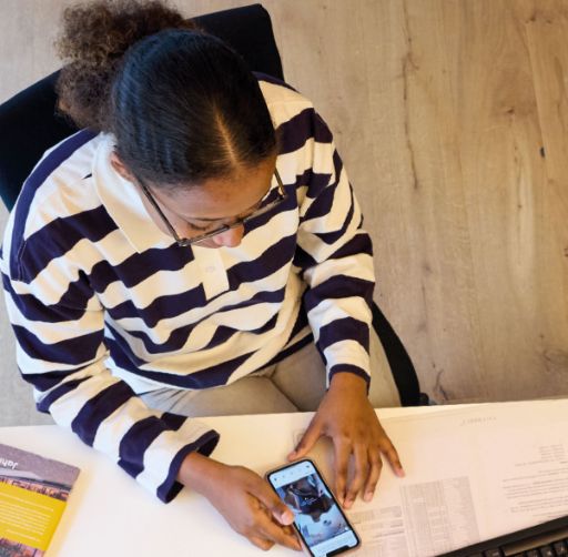 Eine Person mit gestreiftem Pullover sitzt an einem Schreibtisch und betrachtet konzentriert ihr Smartphone, auf dem ein Social-Media-Beitrag zu sehen ist. Auf dem Tisch liegen Papierunterlagen und eine Broschüre, der Hintergrund besteht aus hellem Holzboden.