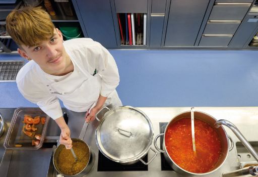 Ein junger Koch in weissem Arbeitsgewand rührt in einem Topf mit gelber Suppe und blickt dabei freundlich in die Kamera. Neben ihm stehen ein weiterer Topf mit roter Gemüsesuppe und eine Schale mit geschnittenen Tomaten auf einer Edelstahl-Arbeitsfläche.