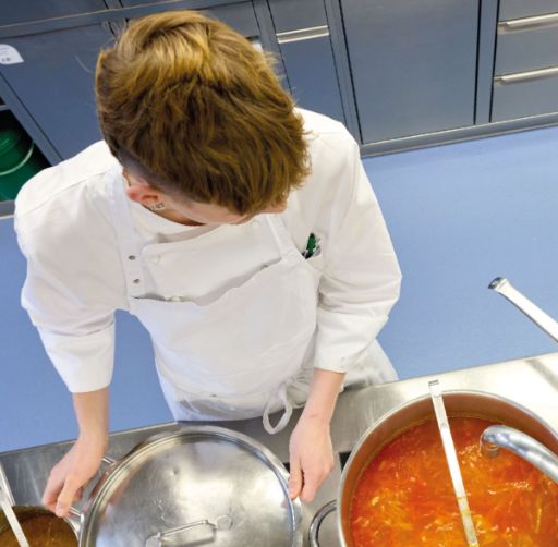 Eine Person in weissem Kochgewand steht in einer Grossküche und hebt den Deckel eines grossen Kochtopfs an. Im danebenstehenden Topf köchelt eine leuchtend rote Suppe.