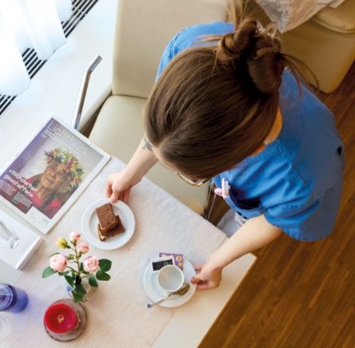Eine Pflegefachperson in blauer Berufskleidung stellt ein Stück Kuchen und eine Tasse Kaffee auf einen gedeckten Tisch. Auf dem Tisch liegen eine Zeitschrift, eine rote Kerze, rosa Rosen in einer Vase und ein Glas Wasser.