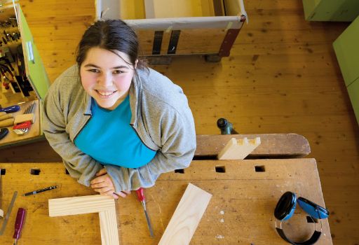 Eine junge Frau in einer Werkstatt steht an einer Werkbank und lächelt in die Kamera, umgeben von Holzteilen und Werkzeugen. Auf dem Tisch liegen unter anderem eine Holzverbindung, Schraubenzieher, Säge und ein Gehörschutz.