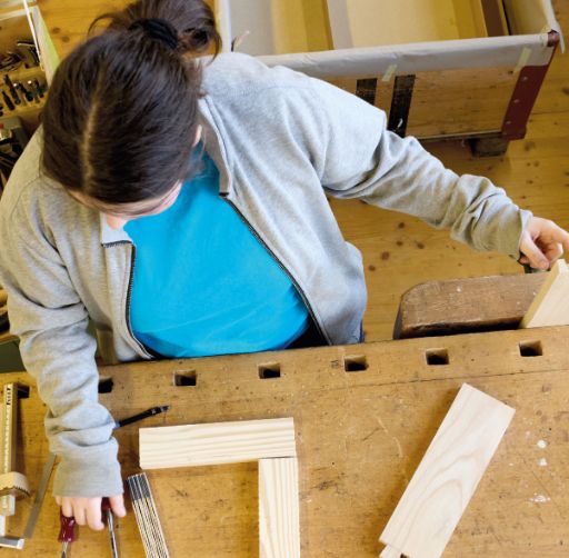 Eine junge Frau in blauem T-Shirt und grauer Jacke arbeitet konzentriert an einer Werkbank und richtet ein Holzstück in einem Schraubstock aus. Auf dem Tisch liegen weitere Holzteile, Werkzeuge und Messhilfen für die Holzbearbeitung.
