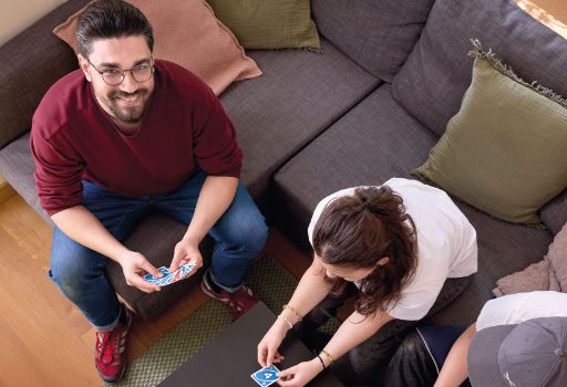 Drei Personen sitzen auf einem Sofa und spielen gemeinsam ein Kartenspiel, wobei eine der Spielerinnen gerade eine Karte auf den Tisch legt. Die Atmosphäre wirkt entspannt und freundlich, einer der Teilnehmenden blickt lachend in die Kamera.