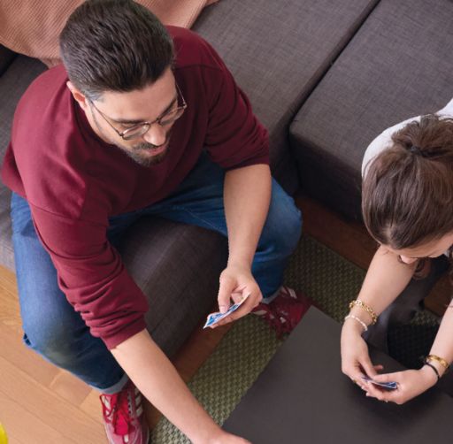 Zwei Personen sitzen auf einem Sofa und spielen ein Kartenspiel, wobei eine Karte gerade auf den Tisch gelegt wird. Beide wirken konzentriert und sind in das Spiel vertieft, die Szene vermittelt eine gemütliche Atmosphäre.