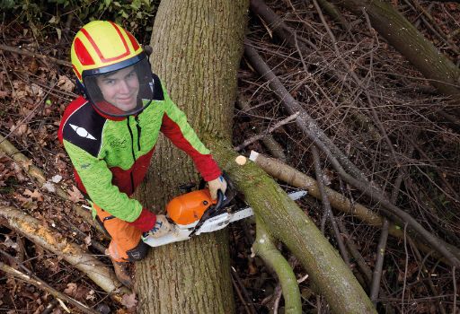 Ein junger Forstwartlernender in auffälliger Schutzkleidung und Helm arbeitet mit einer Motorsäge an einem gefällten Baumstamm im Wald. Um sie herum liegen Äste und Holzreste, der Blick geht freundlich zur Kamera.