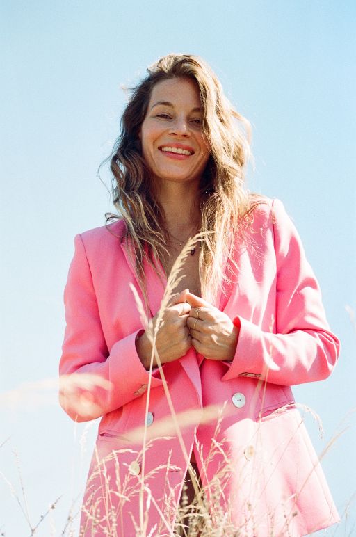 Portrait von Anna Rossinelli vor hellblauem Himmel. Sie lächelt selbstbewusst in die Kamera und trägt einen auffälligen pinken Blazer.