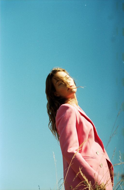 Portrait von Anna Rossinelli vor hellblauem Himmel. Sie ist seiitlich im Bild und schaut über ihre rechte Schulter in die Kamera und trägt einen auffälligen pinken Blazer.