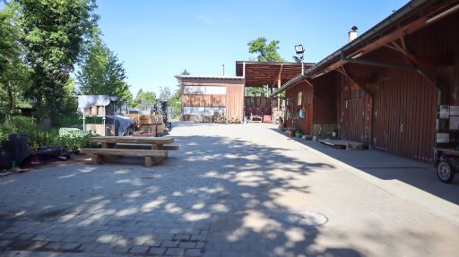 Forstwerkhof der BG Basel mit Holzbauten und gestapeltem Material und Holz. Im Vordergrund stehen ein Holztisch und Pflanzen, im Hintergrund das Hauptgebäude des Forstwerkhofs, Lagerräume und Geräte.