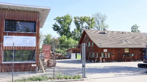 Forstwerkhof der BG Basel mit mehreren Holzbauten und eingezäuntem Areal. Im Vordergrund ein Metallzaun, dahinter gestapeltes Holz, Material und ein Innenhof. Links ein modernes Holzgebäude, rechts ein älteres Holzgebäude mit salbeifarbenen Fensterläden und Pflanzen.