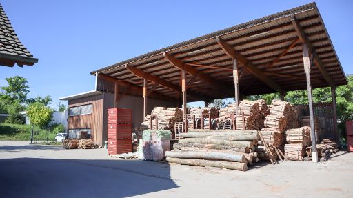 Forstwerkhof der BG Basel mit grossem überdachtem Holzlager und Holzbauten. Im Vordergrund liegen Baumstämme und gestapeltes Holz, daneben kleine rote Container. Rechts davon steht das Betriebsgebäude des Forstbetriebs der BG Basel.