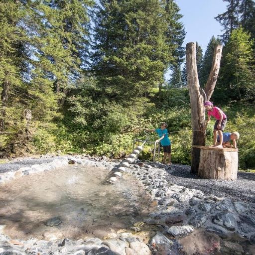 Drei Kinder spielen an einem Wasserspielplatz im Wald mit Steinen und Holzinstallationen. Ein Kind steht auf einem Baumstamm, das andere bedient eine Wasserpumpe am Rand eines kleinen Beckens.