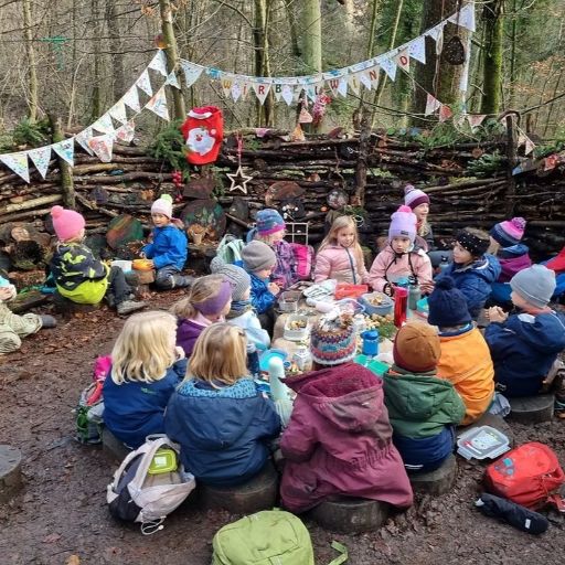 Kinder sitzen im Wald im Kreis und essen gemeinsam Znüni. Hinter ihnen hängt eine Girlande und selbstgemachte Dekoration.