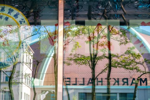 Spiegelung von Bäumen, Fahrrädern und umliegenden Gebäuden in einer grossen Glasfassade. Im Hintergrund ist spiegelverkehrt der Schriftzug «Markthalle» zu erkennen, wodurch ein vielschichtiges Bild aus Architektur, Natur und Reflexionen entsteht.