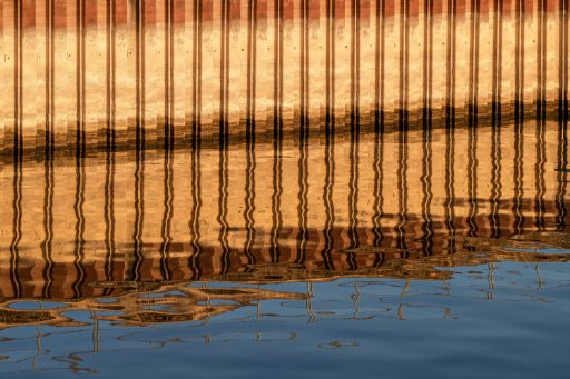 Abstrakte Spiegelung einer goldfarbenen Hafenmauer in Basel mit senkrechten Linien auf einer ruhigen Wasseroberfläche. Die Wellen verzerren das Spiegelbild und erzeugen ein grafisches Muster in Gold, Braun und Blau.